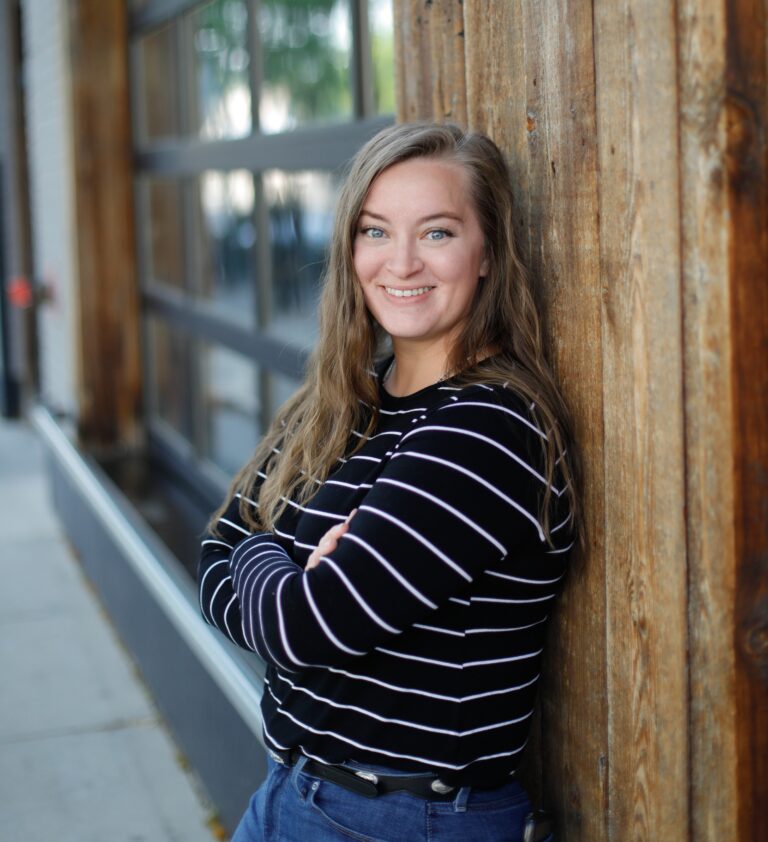 Taylor in a black and white striped shirt, leaning against a wall made of wood. Her arms are crossed and she is smiling.